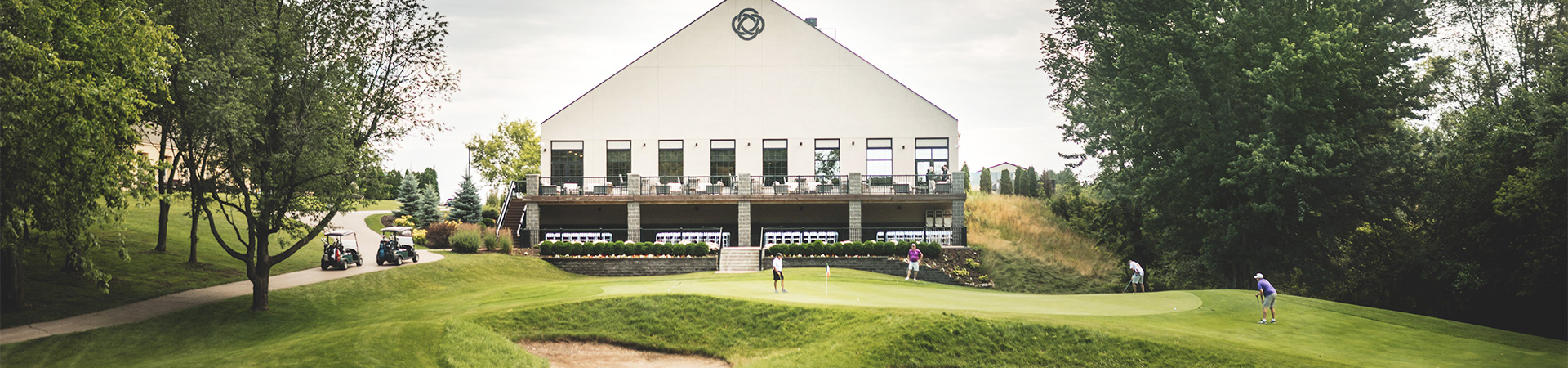 Golfers playing on a green with a clubhouse in the background, surrounded by trees and a well-maintained landscape.