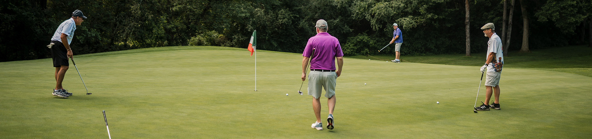 Four golfers on a putting green, focusing on their shots with a flag in the background.