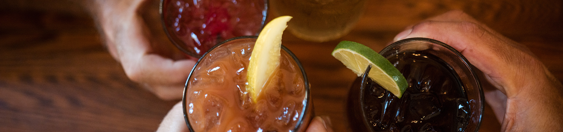 Four hands holding glasses of colorful drinks with ice, garnished with lemon and lime slices.
