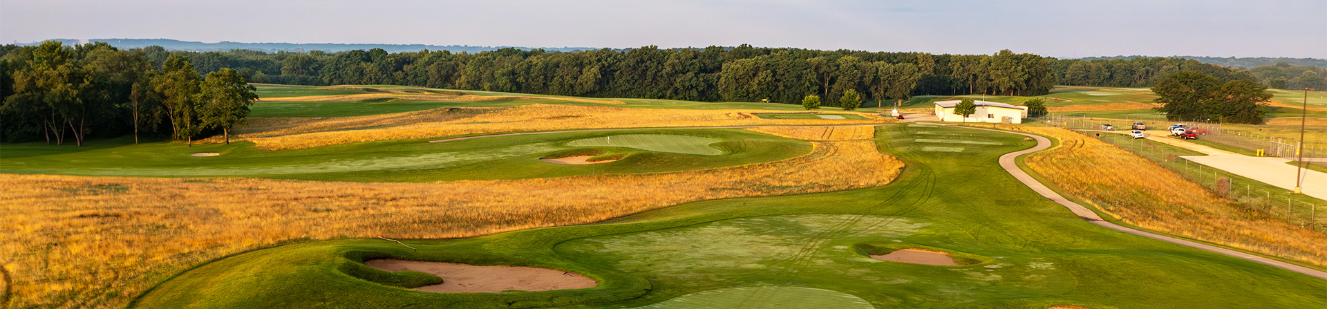 Aerial view of a golf course with green fairways, sand traps, and a clubhouse surrounded by trees and open fields.