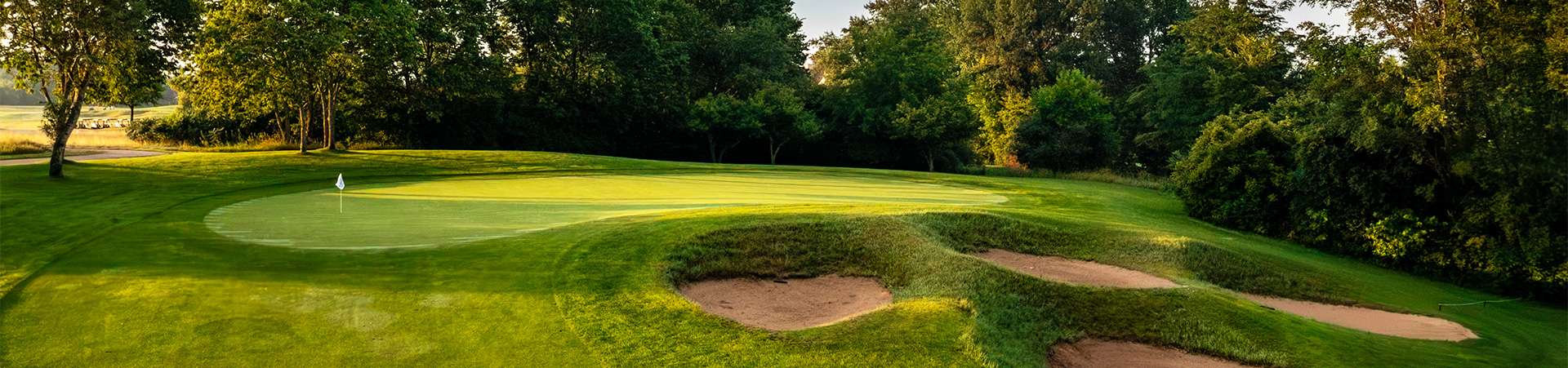 A scenic golf course with a green, sand traps, and trees under soft sunlight.