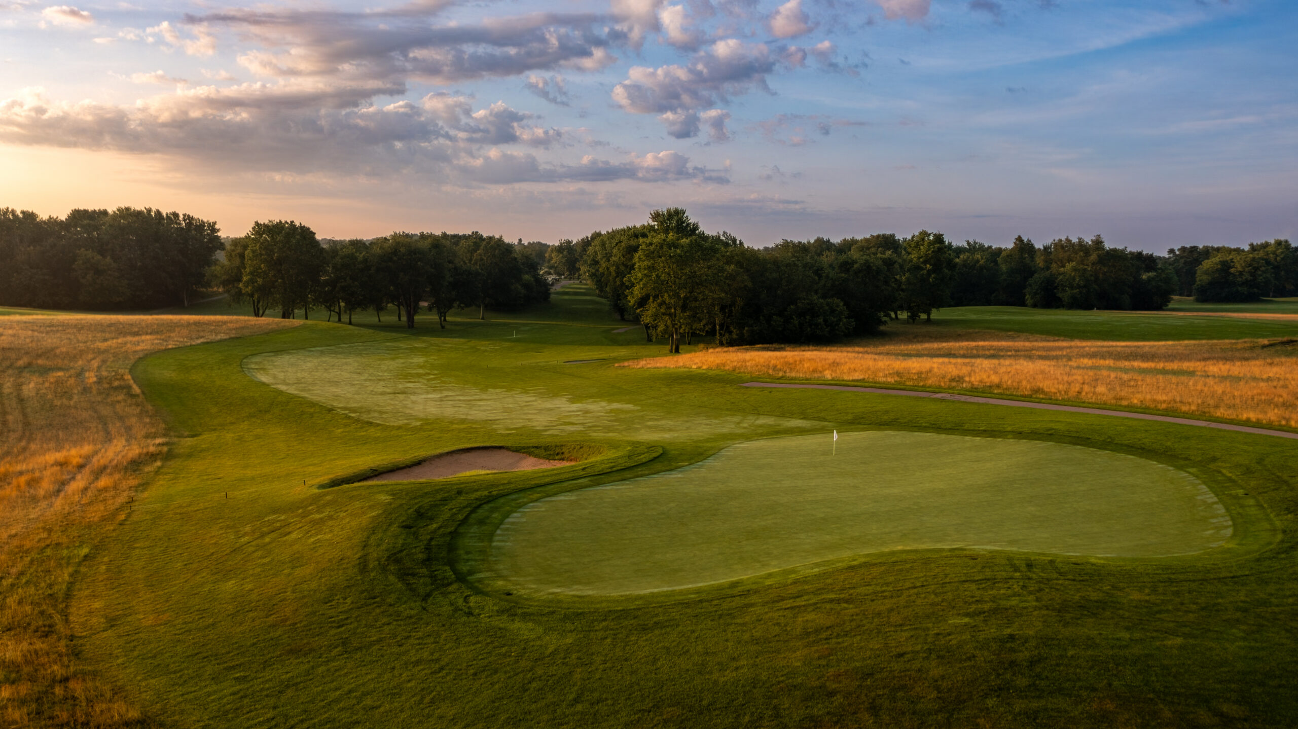 A scenic view of a golf course hole with a green, sand trap, and trees under a cloudy sky at sunset.