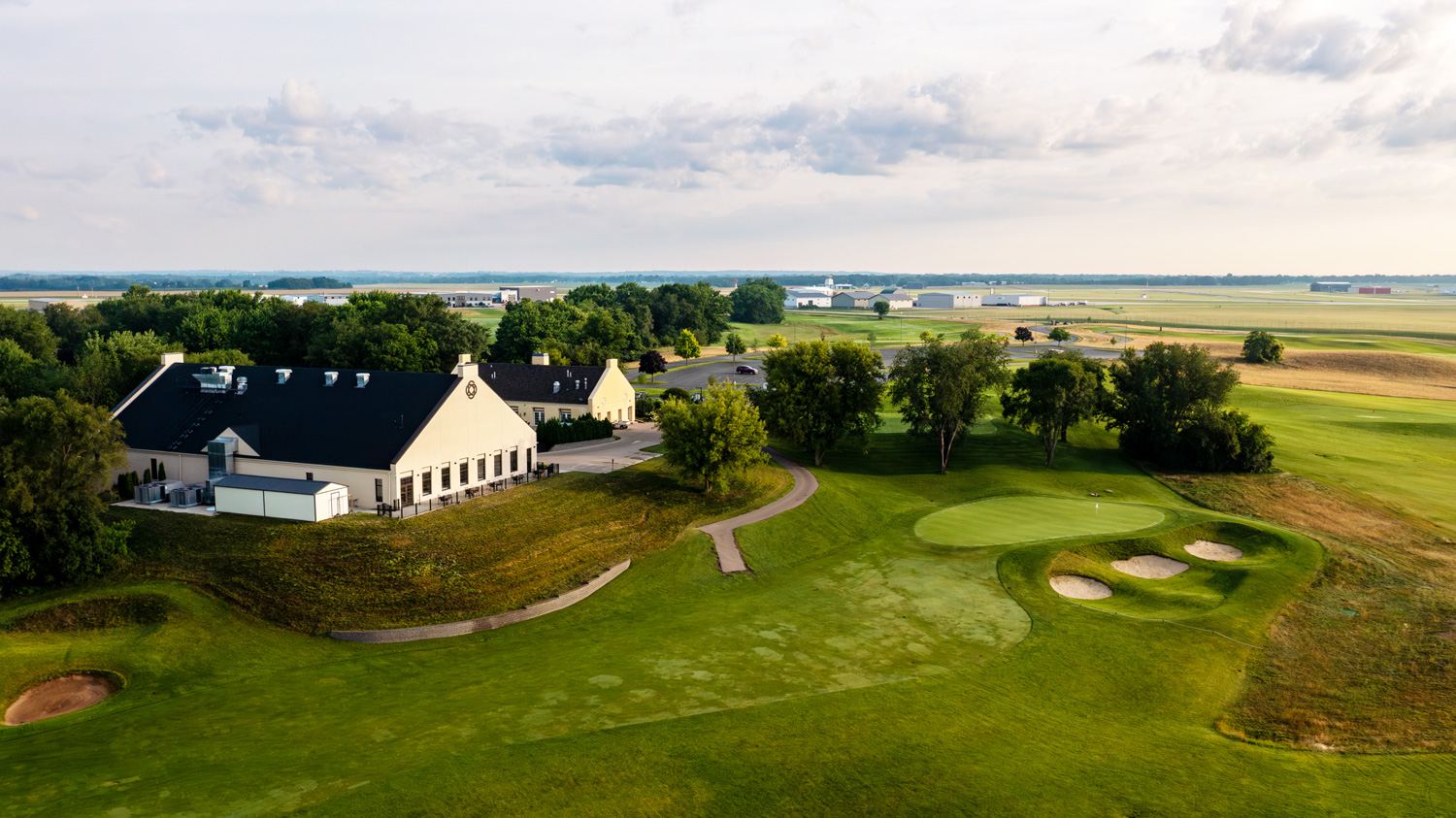 Aerial view of a golf course with a clubhouse, sand traps, and lush green fairways under a cloudy sky.